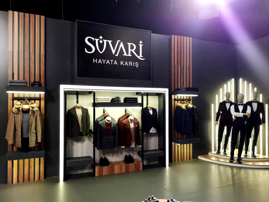 Suvari Logo on a large background of a Expo Stand. Predominant matte black background with wood panels in between men clothing of illuminated suits.