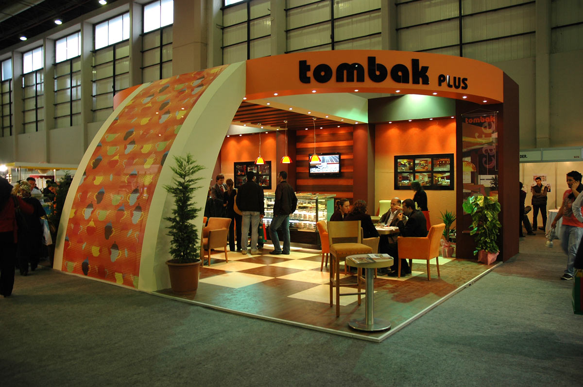 A few people sitting in the Tombak plus large Bespoke orange Exhibition Stand. Has orange brown and cream seating furnitures.