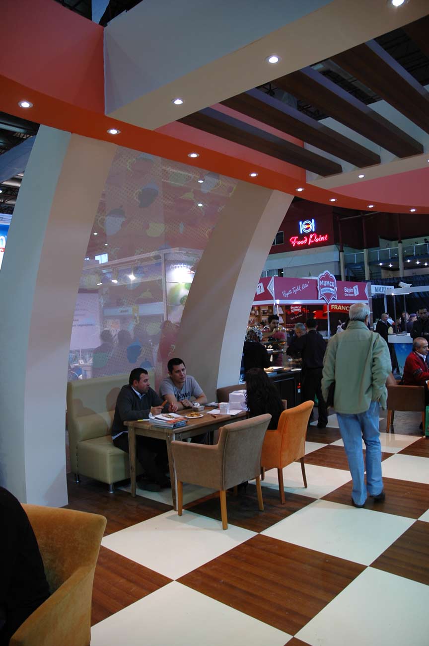 Open seating area with two people sitting and eating sweets in an Exhibition booth in Istanbul. Chessboard tiles on the floor in brown and white.
