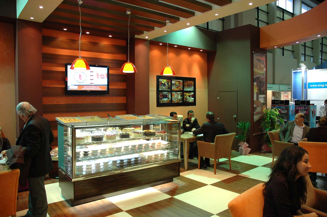 A cafe with modern decor, including striped walls and ceiling, chessboard tiles on the floor. Large transparent fridge in the middle under three lights.