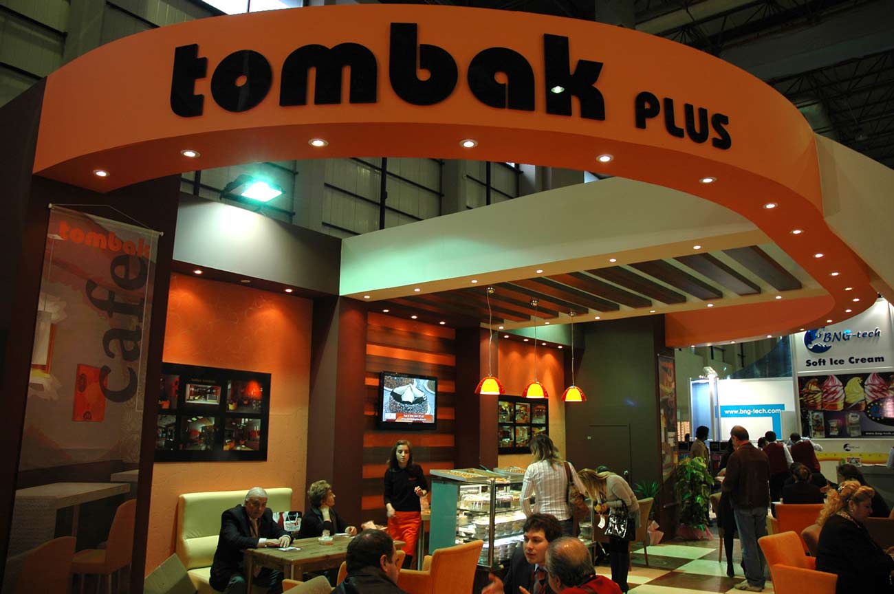 Large black Tombak plus sign on orange background above the Bespoke Exhibition Stand with people under sitting and eating in the cafe.