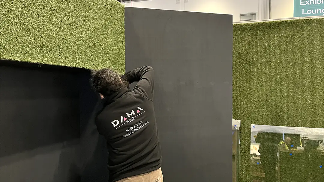 man in dark fleece written Dama design building a grey wall that has grass on it